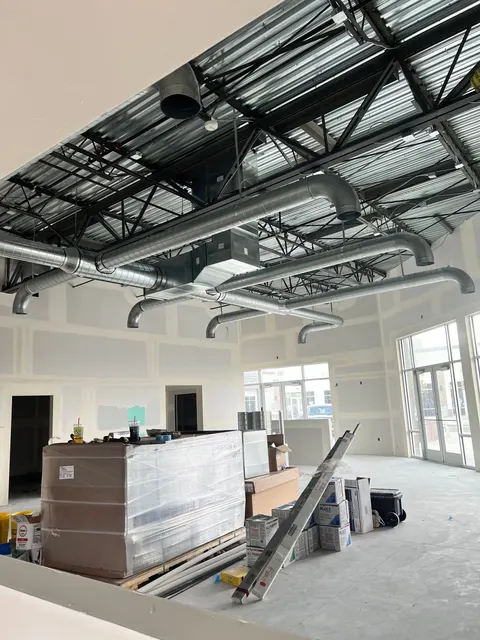 A commercial interior under construction with exposed steel roof framing and ductwork, unfinished drywall, and stacked materials.