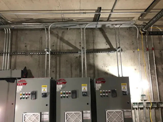 Gray electrical control cabinets mounted against a concrete wall beneath rows of metal conduit.