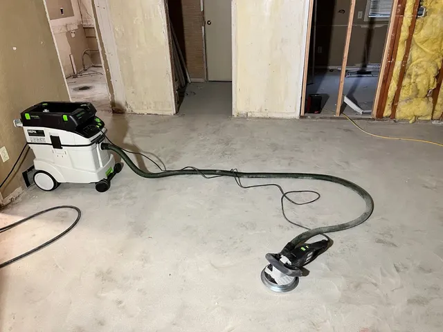 A floor-preparation tool connected to a dust extractor on a bare interior floor, with exposed wall framing and insulation nearby.