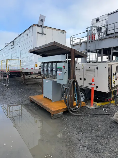 A steel-framed temporary power distribution assembly with electrical enclosures and coiled cables beside a generator and worksite trailer.