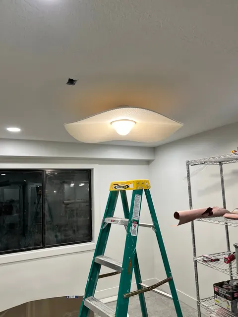 An illuminated sculptural ceiling fixture above a ladder in an interior room with shelving and a window.